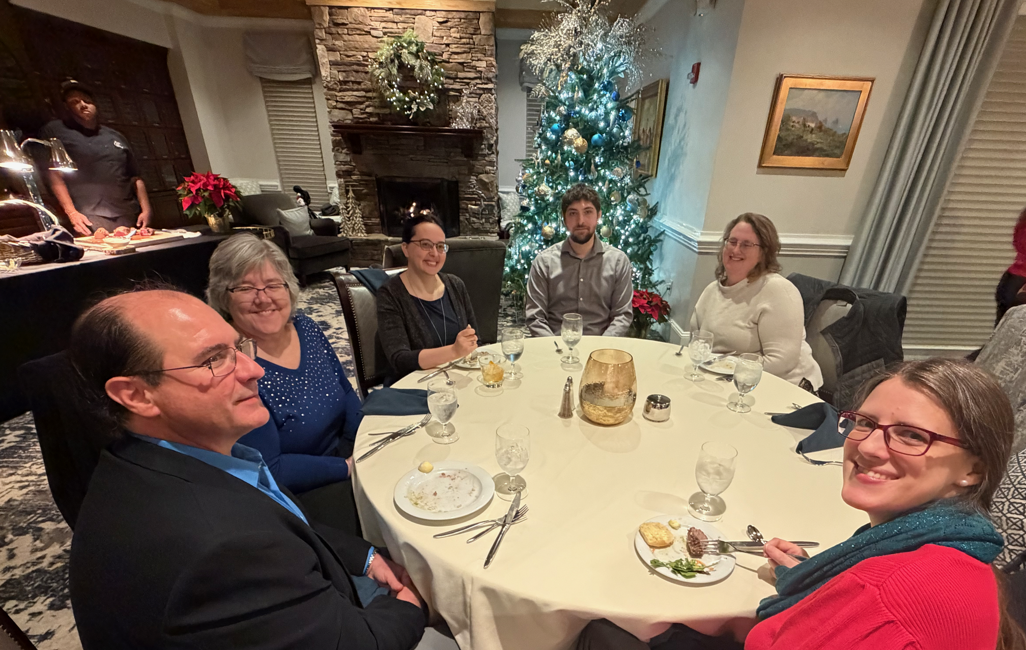 At the next table are Kyle, Nancy, Katy, George, Marion, and Katharina. A beautifully decorated Christmas tree is in the background near a cozy fireplace.