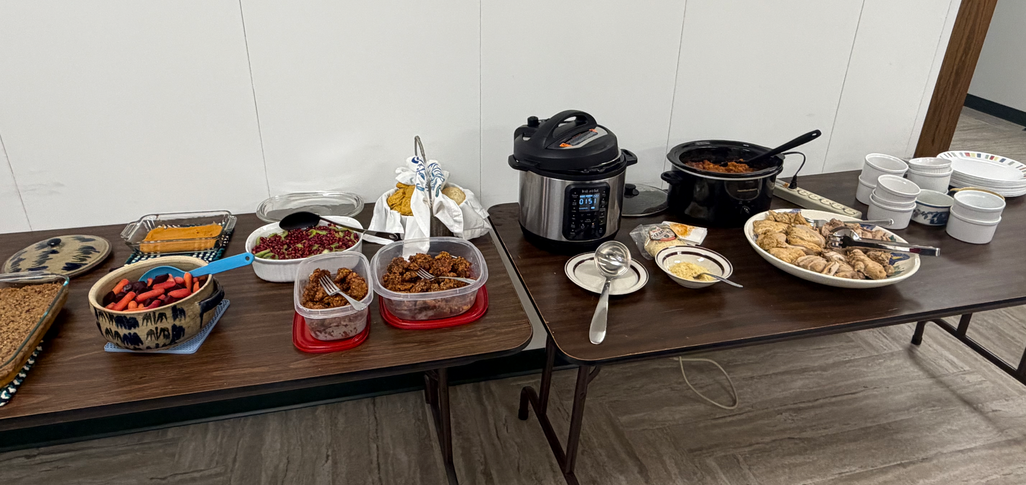 Two typical office tables, loaded with potluck dishes. Offerings included chicken, meatballs, roast veggies, sweet potato casserole, rolls, and a fruit salad with kiwi and pomegranate. There is also a pressure cooker on the table.