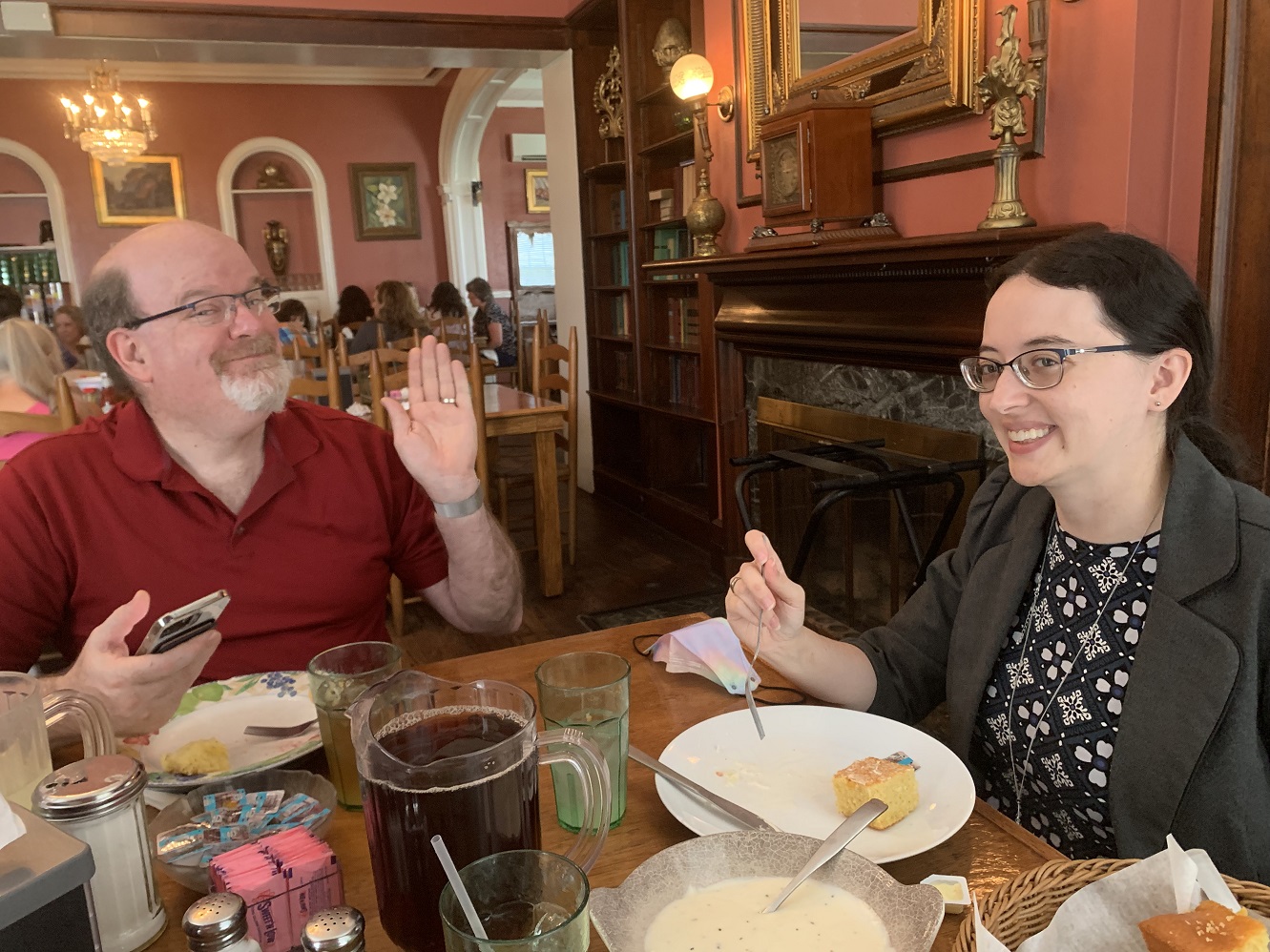 Katy and James smiling at a restaurant table. James is waving to the camera again, and both have plates in front of them with biscuits and cornbread. There is also ice tea and a white gravy visible on the table. The background shows the restaurant's historical architectural features, including a marble fireplace, built-in shelves, and chandeliers, plus eclectic antique décor.