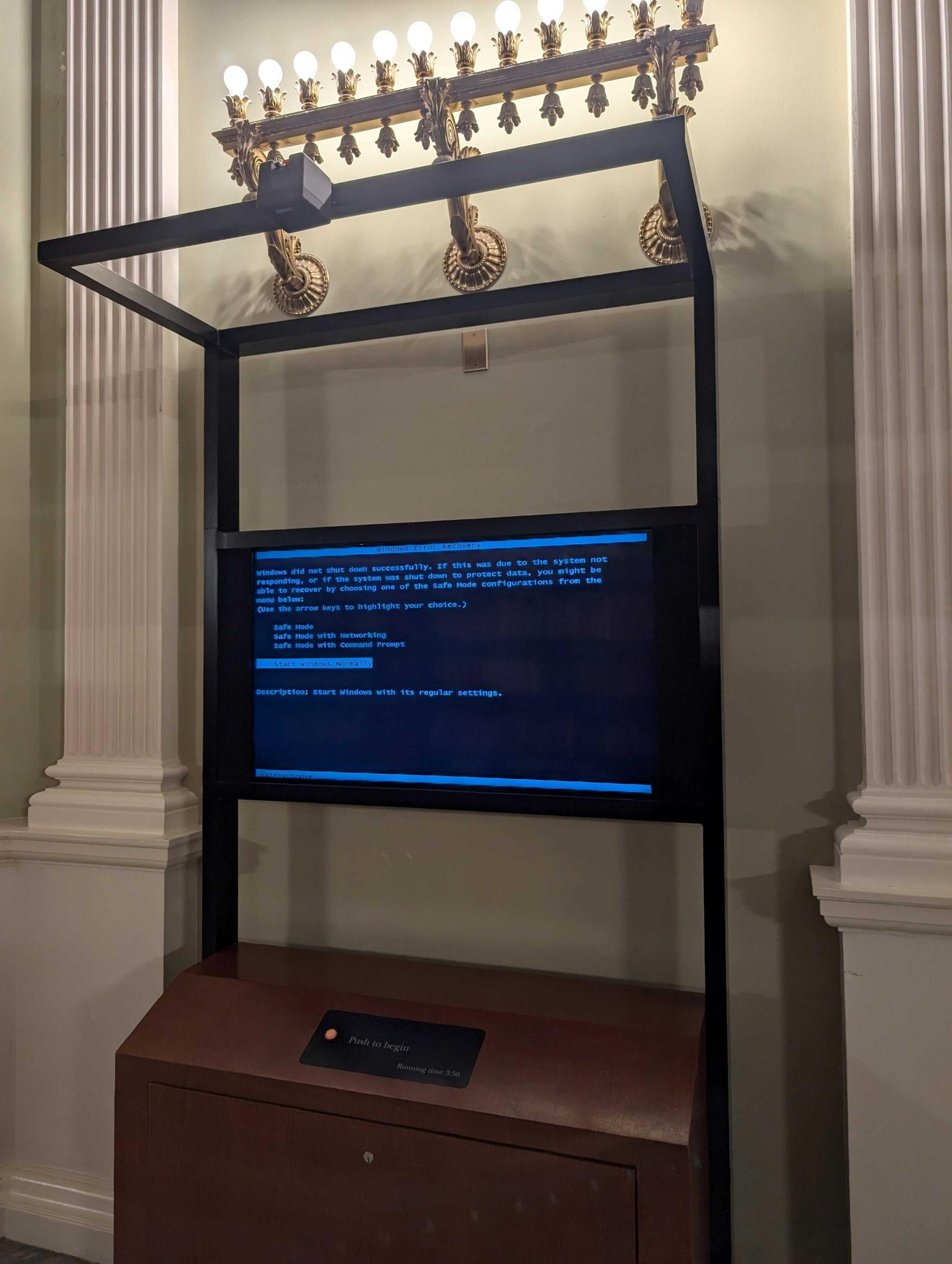 A display stand supports a large screen, with a classic "blue screen" windows error page. Overhead is a fancy, antique sconce lighting the display.